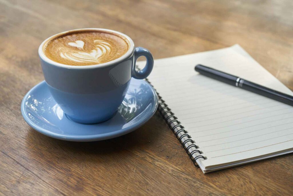 Taza de café en una mesa junto a un cuaderno y un bolígrafo, representando la escritura creativa.