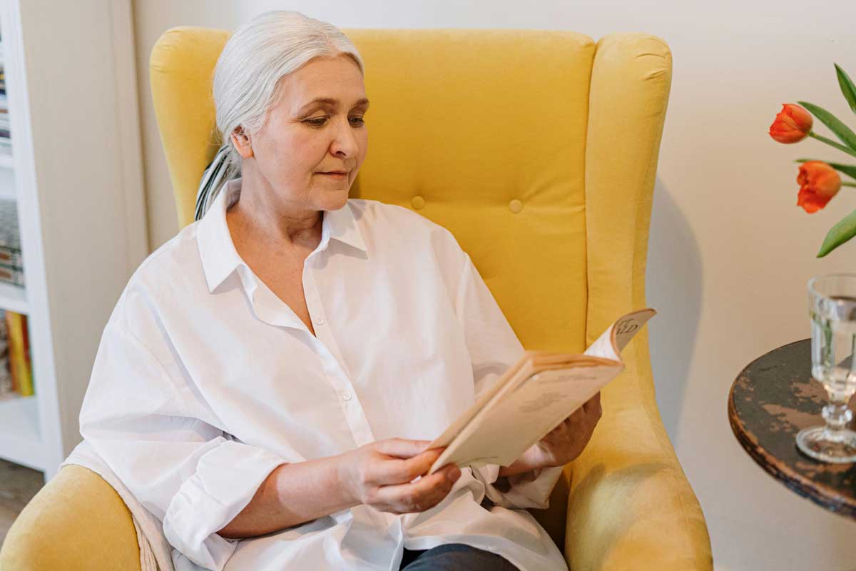 Mujer adulta mayor leyendo un libro en un sillón amarillo, rodeada de un ambiente tranquilo con flores y un vaso de agua.